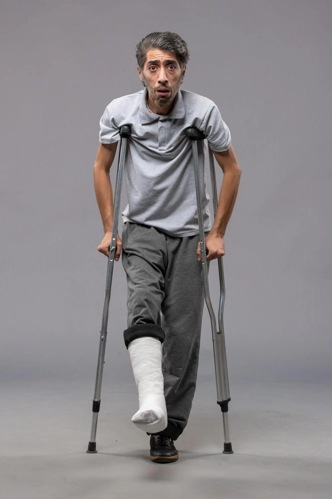 Front view of a young male using crutches due to a broken foot, in front of a grey wall, in pain, with a disabled leg from an accident.