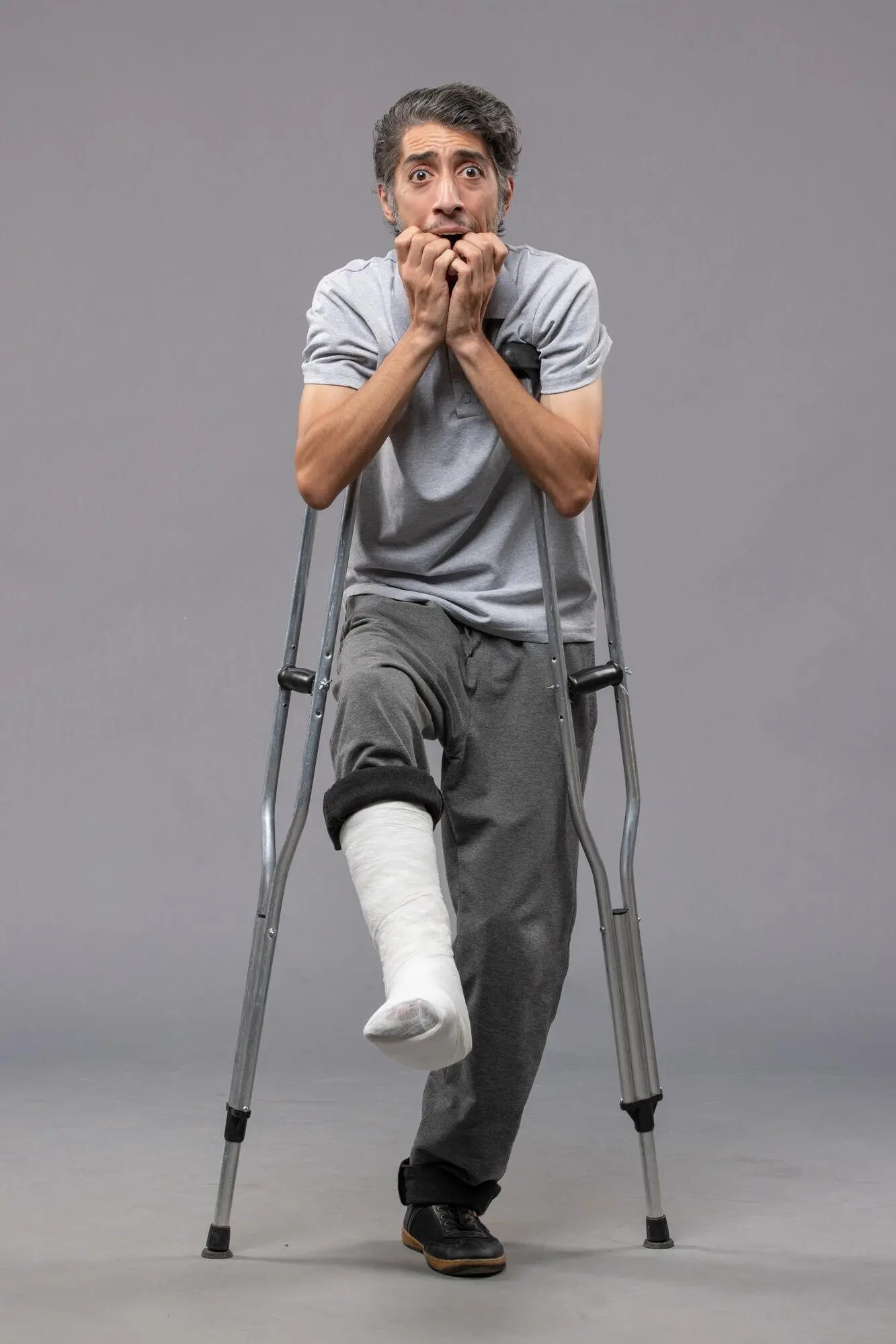 Front view of a young man using crutches because of a broken foot, against a gray wall; disabled; accident-related broken leg/foot.